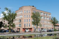 Een gebouw met vijf verdiepingen op een hoek van een straat aan een kanaal met veel waterlelies.