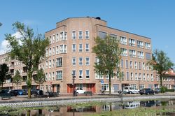 Een gebouw met vijf verdiepingen op een hoek van een straat aan een kanaal met veel waterlelies.
