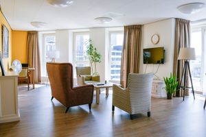 Een lichte woonkamer met veel ramen en een gele muur, staan 3 fauteuils, grote planten en een grote lamp. In de hoek staat een tafel met stoel. Aan de muur hangt een televisie.