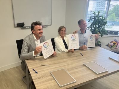 Drie mensen in nette kleding zitten achter een houten tafel en houden blij een contract omhoog.