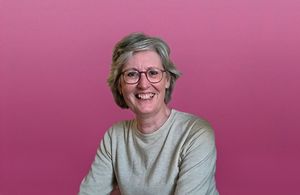 Een portret van een vrolijke vrouw met kort haar en een bril, voor een roze achtergrond.