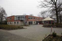 Een gebouw met twee verdiepingen met een metalen trap naar de tweede verdieping. Rechts aan het gebouw zit een rond bijgebouw met wit puntdak.