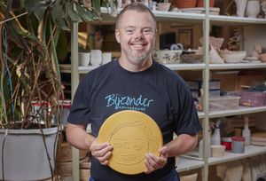 Een man staat blij geposeerd voor een kast met allerlei keramieken producten. In zijn hand heeft hij een keramieken bord en hij draagt een shirt met de tekst Bijzonder Amsterdams erop.