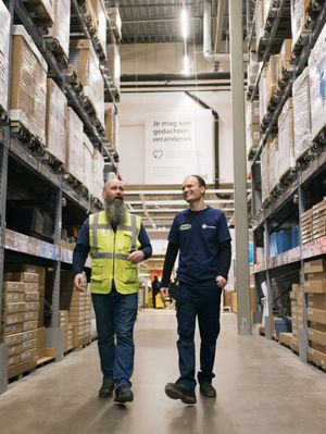Twee mannen lopen lachend door een magazijn. De rechter heeft een lange baard en draagt een geel hesje en de linker heeft een blauw shirt aan met Cordaan-logo.
