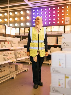 Een persoon met hoofddoek en geel hesje staat lachend op de lampenafdeling van een winkel.