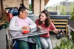 Een vrouw met donker haar en een meisje in een rolstoel kijken op een groot balkon naar de planten. De vrouw heeft een plantenspuit vast.