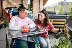 Een vrouw met donker haar en een meisje in een rolstoel kijken op een groot balkon naar de planten. De vrouw heeft een plantenspuit vast.
