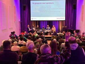 Een sfeervol verlichte zaal met mensen die naar een podium kijken met verschillende sprekers op stoelen en daarachter een groot scherm met hun namen erop.