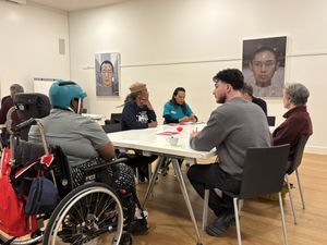 Een witte ruimte met daarin een tafel met daaraan zes mensen in gesprek. Een van hen zit in een rolstoel. Aan de muur hangen portretfoto's.