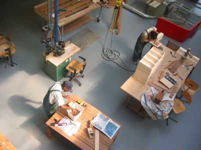 Bovenaanzicht van de houtwerkplaats. Er zijn verschillende werkbanken en grote apparatuur zichtbaar. Twee mensen in groene overals werken aan houten meubels.