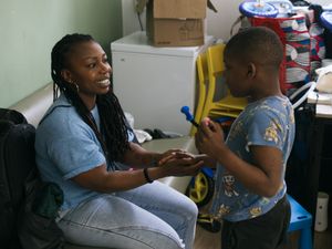 Een vrouw met donker, ingevlochten haar zit op een bank en houdt de hand vast van een jongetje.