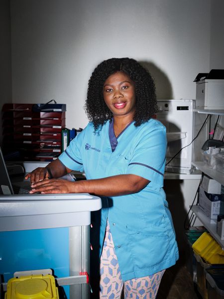 Een vrouw met zwarte krullen staat in een medicijnkast. Ze draagt een lichtblauw werkuniform met Cordaan erop.