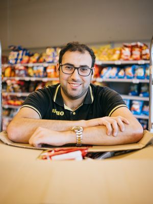 Een man met donker haar, een bril en een zwarte polo met de gele letters Jumbo erop staat voor een supermarkt schap met chips. Hij leunt op een kartonnen doos en kijkt lachend in de camera.