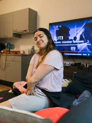 Een kleine, jonge vrouw zit op een bank en kijkt blij in de camera. Op de achtergrond staat de tv aan.