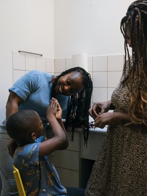 Twee vrouwen met donker, ingevlochten haar staan in de keuken en kijken lachend naar een jongetje op een gele stoel.