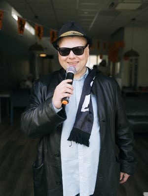 Een man met een zonnebril, zwarte leren jas en hoedje met de naam André Hazes erop zingt in een microfoon.