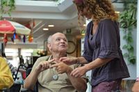 Vriendelijk kijkende oudere heer met zorgalarm om zijn nek doet oefening voor fijne motoriek met zijn handen. Een begeleider met lange bruine krullen staat naast hem en kijkt naar de handen van de mijnheer