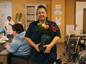 Een medewerkster met donkerblauw Cordaan uniform staat in een gezellige woonkamer met gele muren en houdt een vaas met bloemen vast.