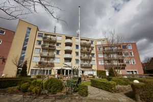 Gebouw uit lichte en rode baksteen opgetrokken, met veel ramen, balkons en een tuin ervoor met haag en stenen pad ertussendoor.