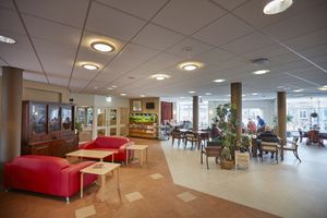 Een grote ruimte met daarin tafels en stoelen met mensen, twee rode banken naast een houten buffetkast en midden in de zaal grote pilaren tot het plafond.