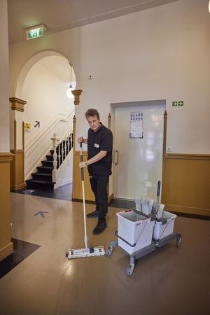 Een man met zwarte kleding staat in de hal van een oud gebouw een maakt de vloer schoon met een vloerwisser.