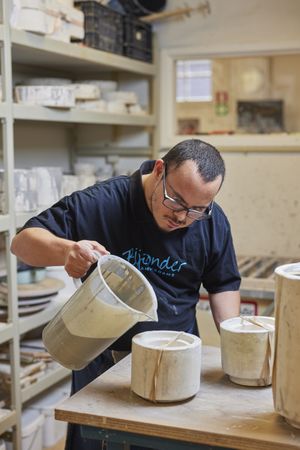 Een man met bril met een zwart shirt met de tekst 'Bijzonder Amsterdams' giet geconcentreerd vloeibare klei in een stenen mal.