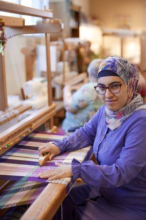 Een vrouw met paarse blouse, hoofddoek met bloemenprint en bril weeft een paarse stof met een weefgetouw en kijkt in de camera..