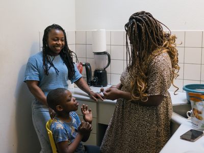 Twee vrouwen met donker, ingevlochten haar staan in de keuken en kijken lachend naar een jongetje op een gele stoel. De ene vrouw geeft het jongetje een snoepje.