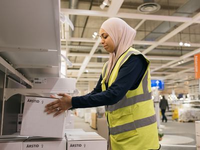 Een persoon met hoofddoek en geel hesje tilt een witte doos in een winkelschap.