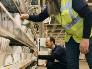 Twee personen vullen de schappen van een winkel met dekbedovertrekken. Ze zitten op hun hurken.