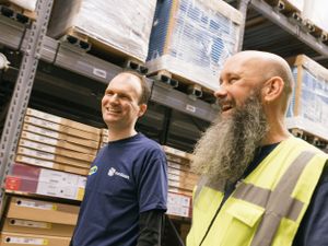 Twee mannen lopen lachend door een magazijn. De linker heeft een blauw shirt aan met Cordaan-logo en de rechter heeft een lange baard en draagt een geel hesje.