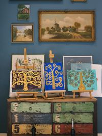 Een oude kast met daarop drie kleine schildersezels met schilderijen van bomen met krullende takken. Daarboven aan de muur drie schilderijen van boerderijen.