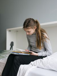 Meisje met grijs shirt en een paardenstaart zit op een wit bed een boek te lezen.