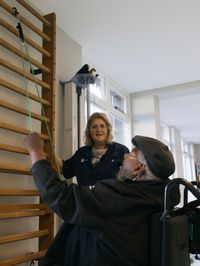 Een man en een vrouw zijn in een sportruimte. De man zit in een rolstoel en trekt aan een touw aan de muur. De vrouw moedigt hem aan.