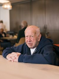 Oudere heer met donkerblauwe hoodie zit aan tafel in het restaurant en kijkt de camera in