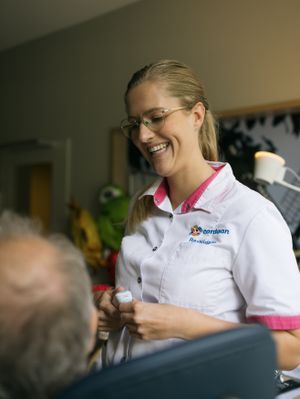 Een vrolijke blonde vrouw met bril lacht naar een man in een stoel. Ze draagt een wit met roze uniform met de tekst 'Cordaan revalidatie' erop.