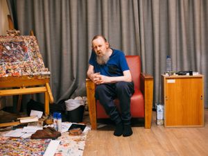 Een oudere man met een lange baard zit op een rode, leren stoel en kijkt in de camera. Naast hem staat een groot schildersezel met een schilderij met heel veel kleine, gekleurde strepen.