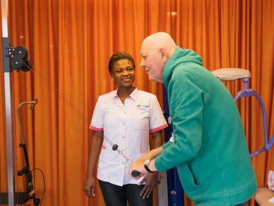 Een vrouw in wit Cordaan uniform kijkt lachend naar een man die oefeningen doet. Hij trekt met beide armen een touw naar beneden.