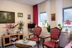 Zitslaapkamer met Lodewijk de veertiende stoelen met rode bekleding, gehaakte kleedjes op tafel, gladiolen in een vaas, een tuinkabouter op een kastje en schilderijen met bosgezicht aan de muur.