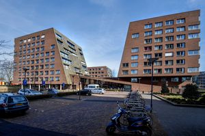 Een groot bruin gebouw met links en rechts een woontoren, verbonden door de begane grond verdieping. Daarvoor een parkeerplaats met auto's en fietsen.