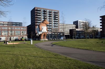 Een flat met meer dan tien verdiepingen en balkons aan een groen park, met voor de flat een imposant beeld van een bruine beer met een kussen in zijn poot.
