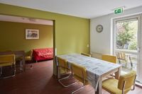 Een met een tafelkleed gedekte eettafel met 5 stoelen met gele ribbekleding. In aansluitende ruimte eenzelfde opzet