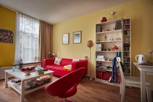 Een appartement met gele wanden, een boekenkast, zithoek met rode stoel en bank en een witte eettafel met legpuzzel erop.