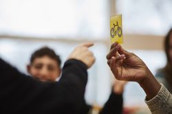 Hand houdt een pictogram omhoog met een fiets erop. Een vinger van andere persoon wijst ernaar, terwijl op de achtergrond iemand lachend toekijkt.