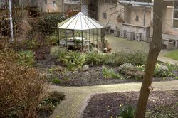 Binnentuin met verharde, paden tussen de planten door. In het midden staat een kas en aan de zijkant staan grote houten tuinmeubels met veel zit plekken.