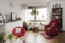 Lichte kamer met grote ramen en een vrolijke rode sta-opstoel, boekenkast, witte dressoir en een tafeltje met gele tulpen