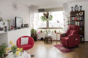 Lichte kamer met grote ramen en een vrolijke rode sta-opstoel, boekenkast, witte dressoir en een tafeltje met gele tulpen