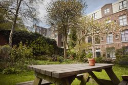 Een tuin met groen gras, bomen in bloei en een tuintafel met bloempotje. De tuin ligt aan een oud uitziend pand.