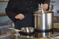 Iemand in zwart koksuniform roert in een grote kookpan op het fornuis.