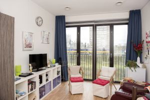 Een kleine kamer met een gezellige sfeer met grote ramen en donkerblauwe gordijnen. In de kamer staan een witte vakkenkast met tv erop en twee rieten stoelen met witte kussens.ls en decoraties.